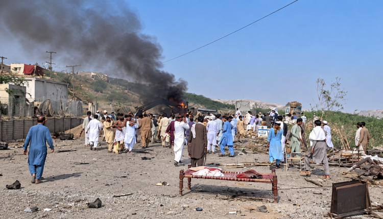 People gather near the site of a suspected suicide blast at an anti-smuggling check post in Dera Ismail Khan’s Darazinda area as smoke rises from the scene.